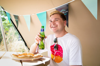 a man holding a bottle of beer smiles with a pizza in front of him