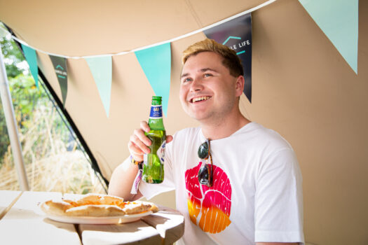 a man holding a bottle of beer smiles with a pizza in front of him